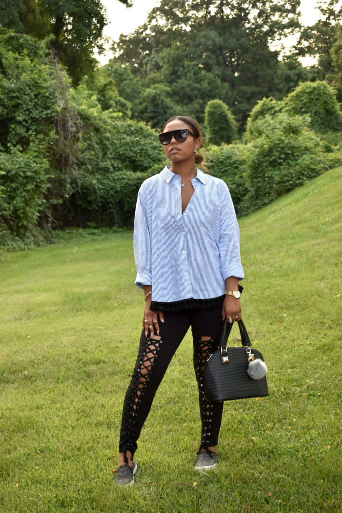 Female standing in front of greenery landscape wearing a blue and white pinstripe button down blouse with black lace up detailed pants and charcoal colored sneakers. She's also holding a black handbag while wearing various pieces of jewelry and a pair of sunglasses.