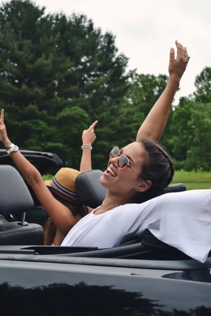 Black convertible with top down and songs playing. Girl sitting in back having fun with friend in passenger seat, both have their arms up.