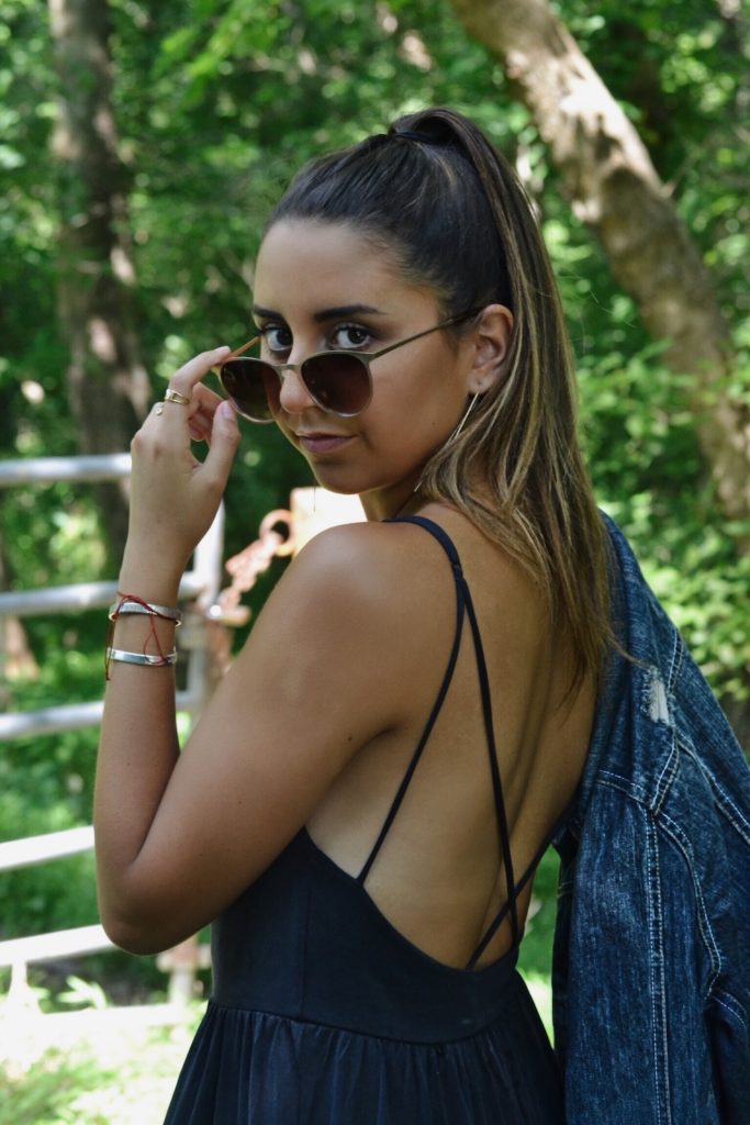 Girl with a high pony hairstyle, wearing culotte jumpsuit peaking over sunglasses with jean jacket hanging over shoulder.