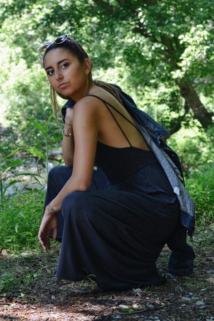 Girl wearing culotte jumpsuit crouched down, with jean jacket thrown over shoulder.